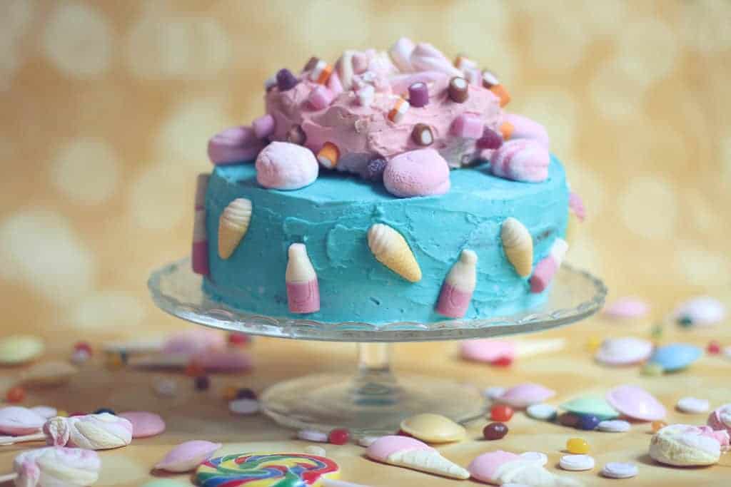 Sponge cake covered with bright blue inning and a pink iced ring on top covered in sweets. Child’s birthday cake.