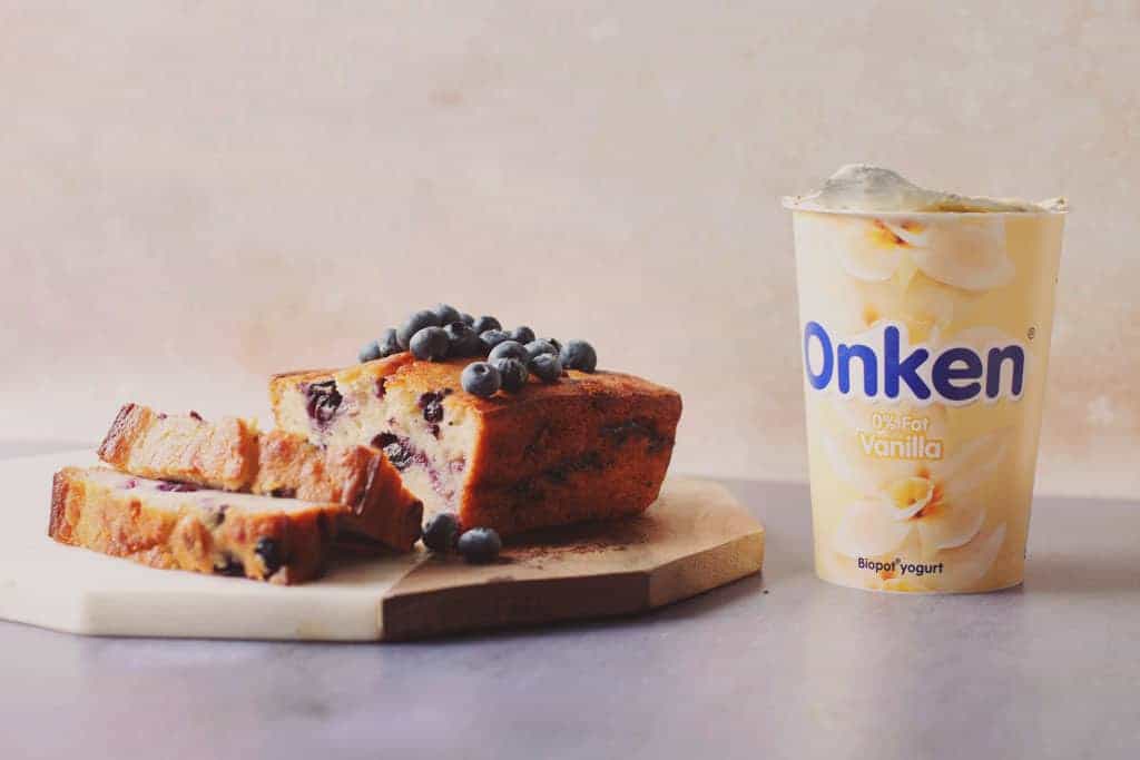 Bluberry banana bread cut into slices and topped with fresh blueberries beside a pot of Onken yoghurt