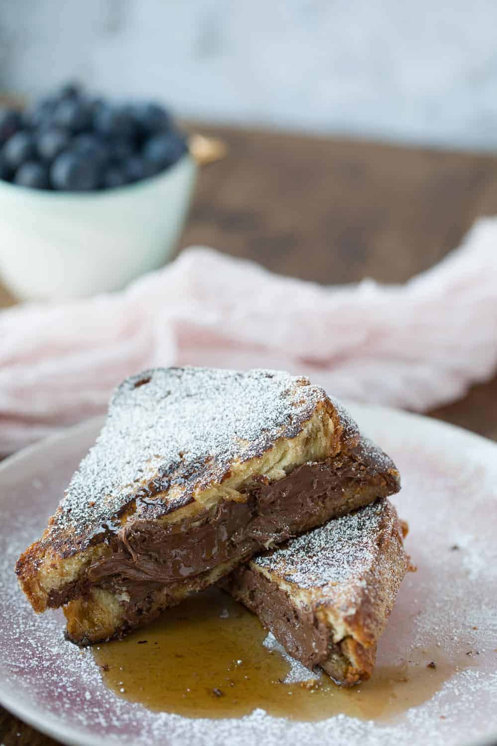 Two triangles of nutella Stuffed French toast on a plate with a bowl of blueberries in the background