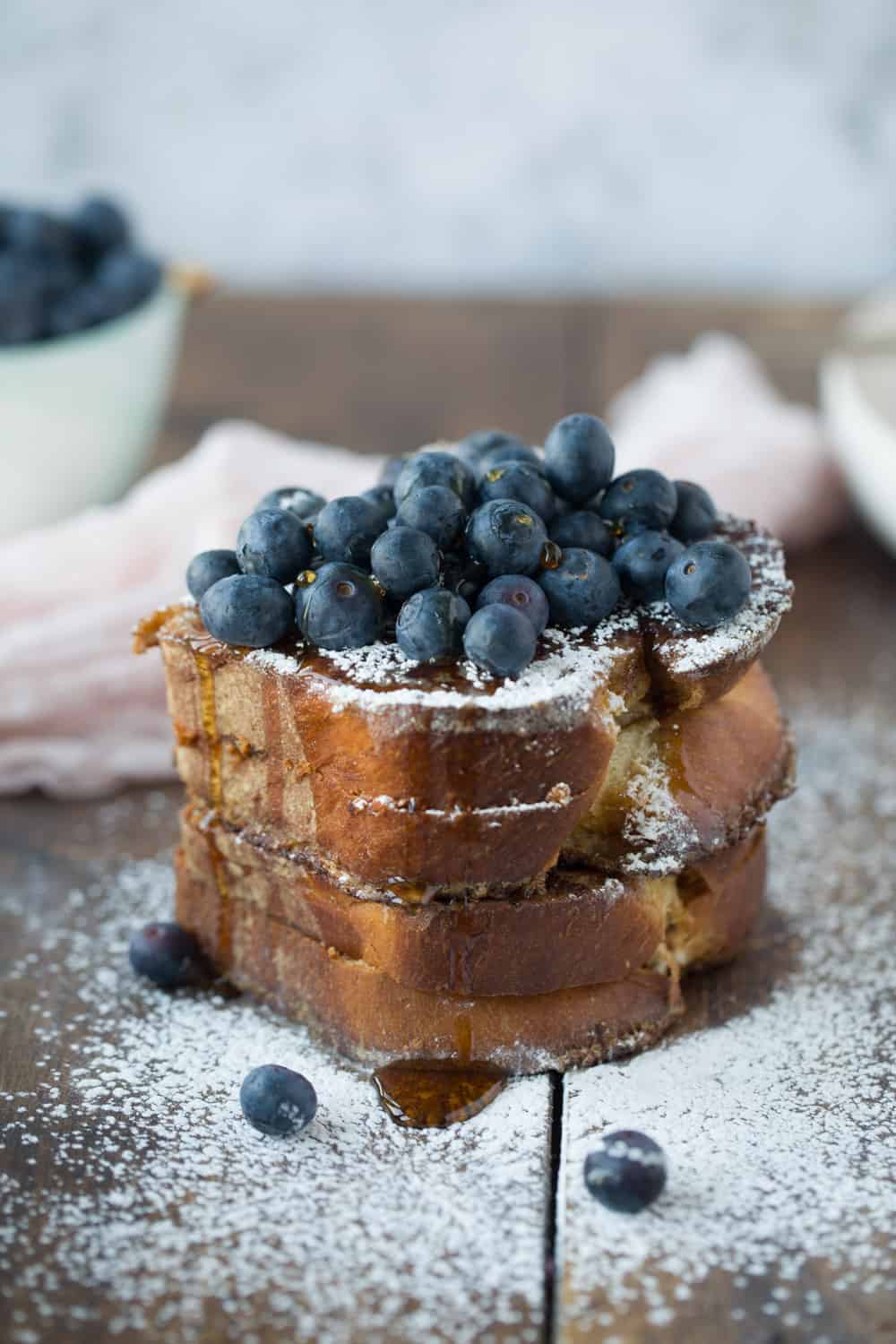 A stack of French toast covered with blueberries, drizzled with maple syrup and sprinkled with powdered sugar