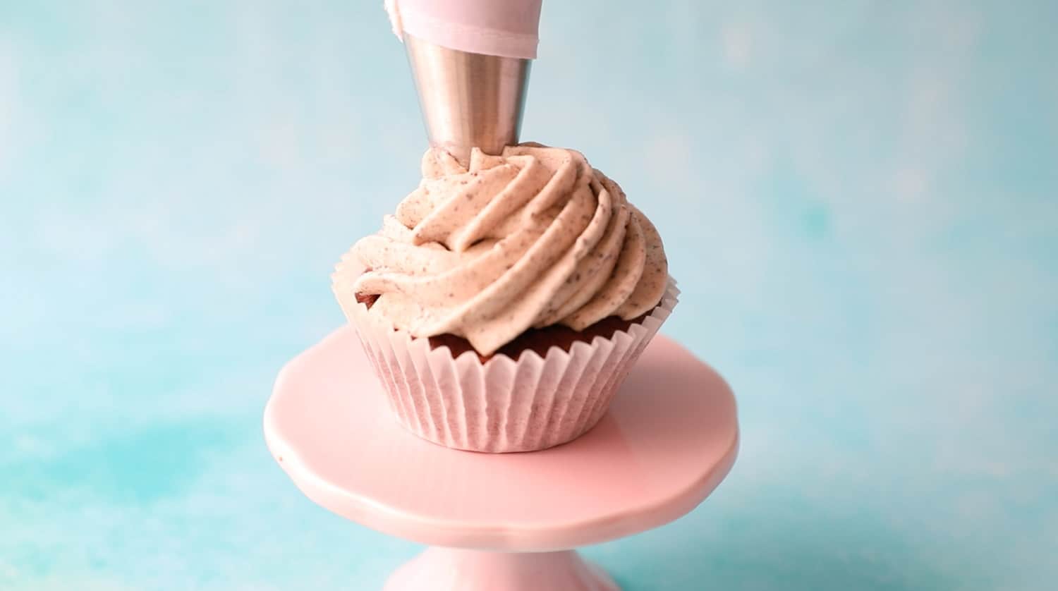 A cupcake on a pink stand with a buttercream swirl also visible is the top of a piping bag fitted with a star tip