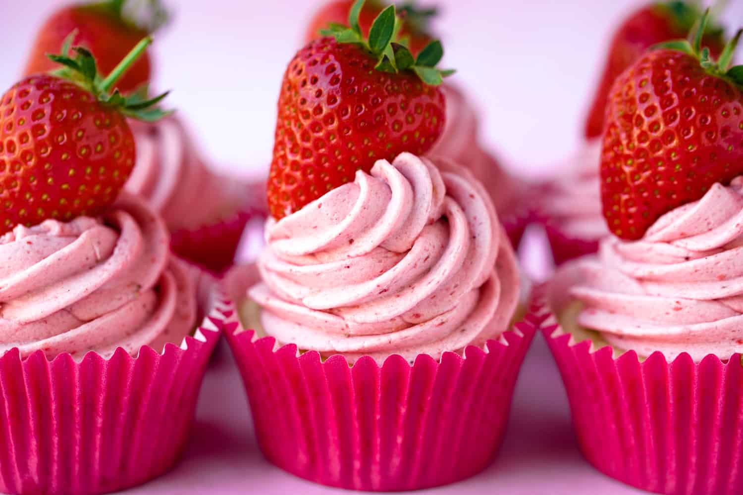 A row of pink strawberry cupcakes in pink cases topped with fresh strawberries
