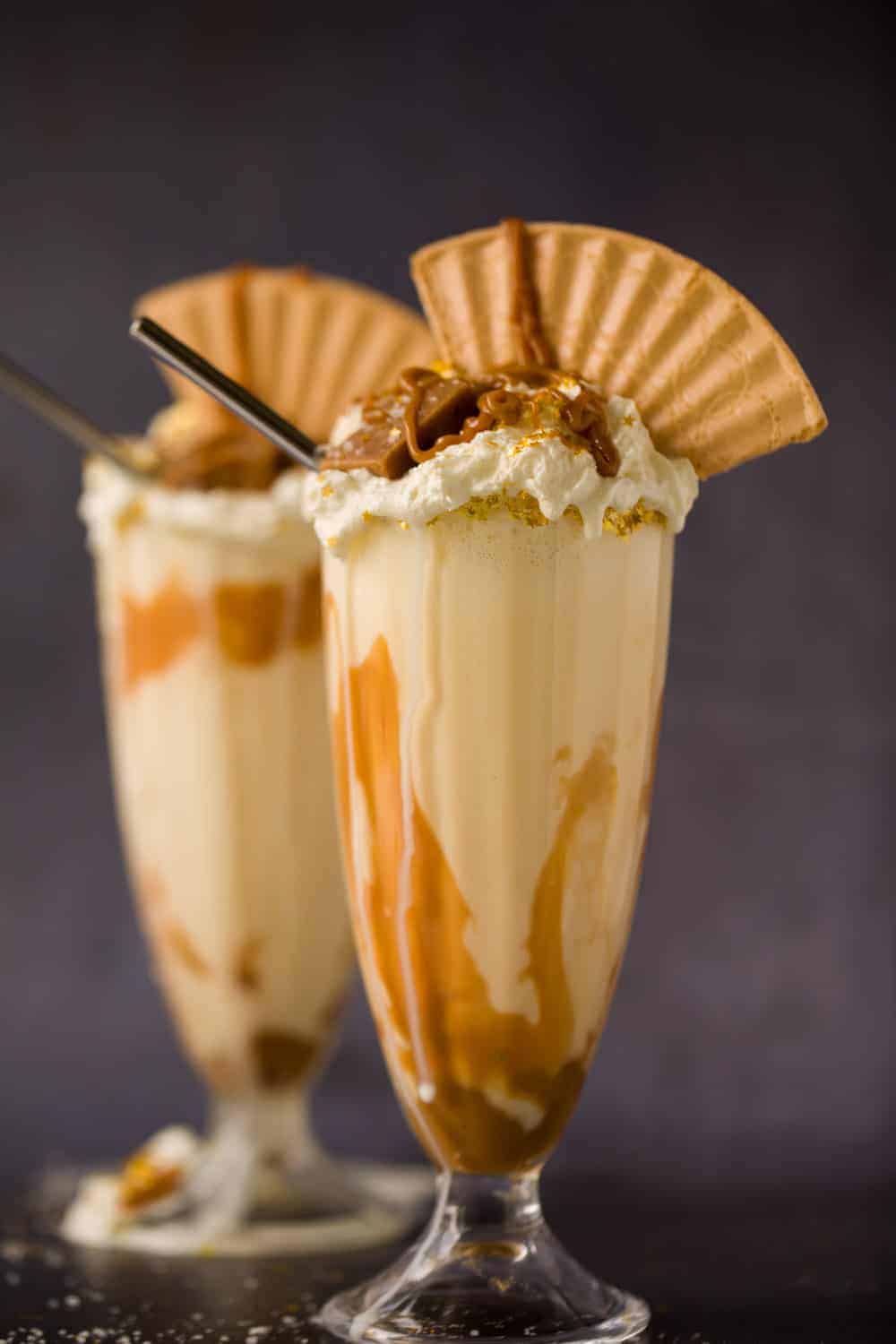 Two tall glasses filled with salted caramel milkshake. The glasses are topped with whipped cream, a wafer and pieces of salted caramel fudge. 