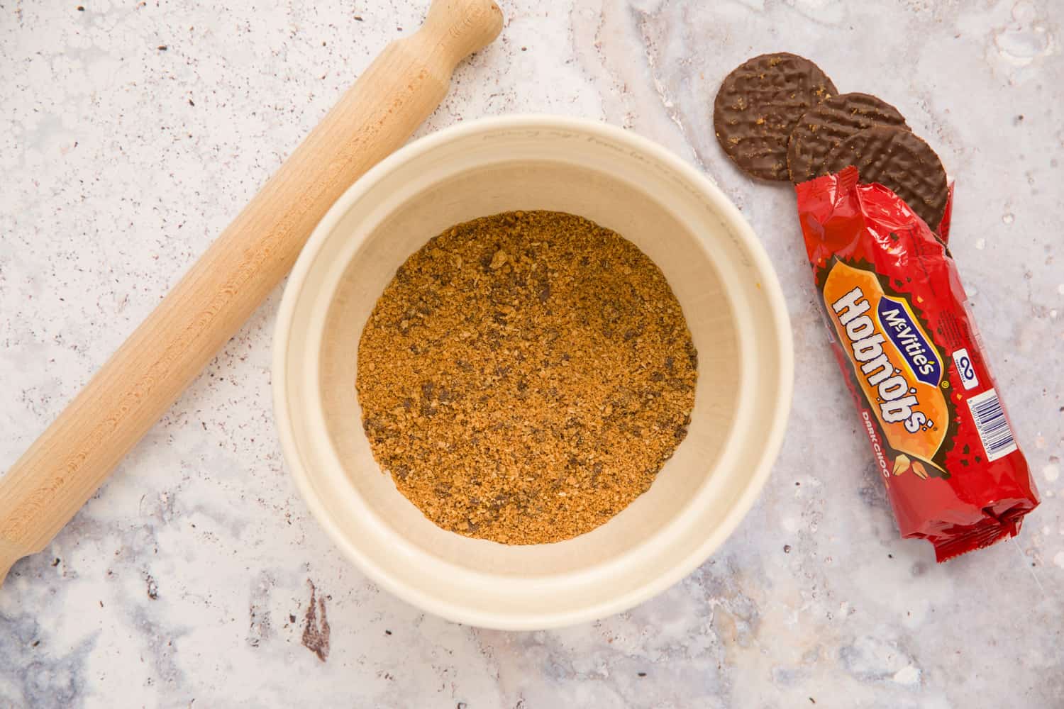 Overhead shot of a mixing bowl filled with crushed Hobnob biscuits. There is a rolling pin to the left of the bowl and a packet of chocolate Hobnob biscuits to the right of the bowl. 