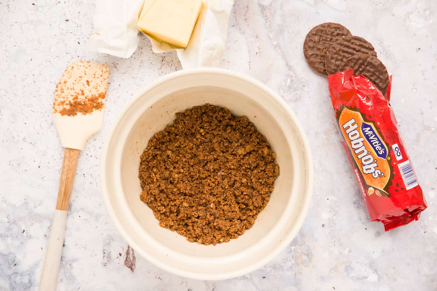 Overhead view of a mixing bowl with crushed biscuits that have been coated in melted butter. There is a spatula to the left of the bowl, above the spatula is some butter. There is a packet of open Hobnob biscuits to the right of the bowl. 