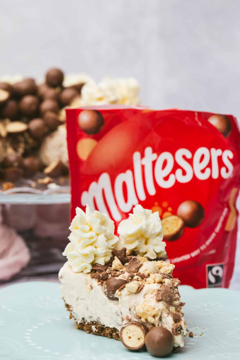 A slice of Malteser cheesecake in the foreground and a bag of red Maltesers in the background