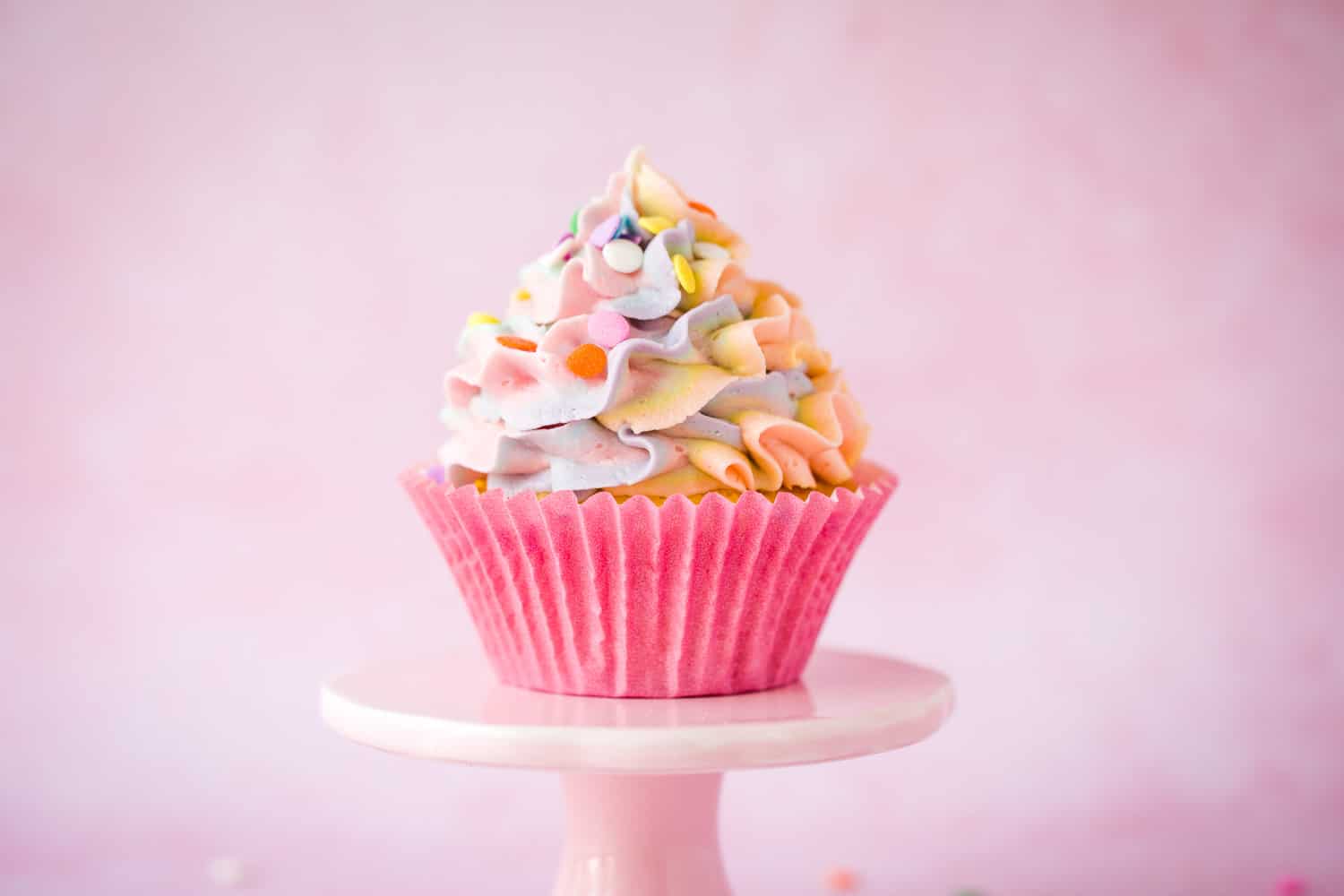 A piñata cupcake on a small pink cake stand with ruffle rainbow coloured icing