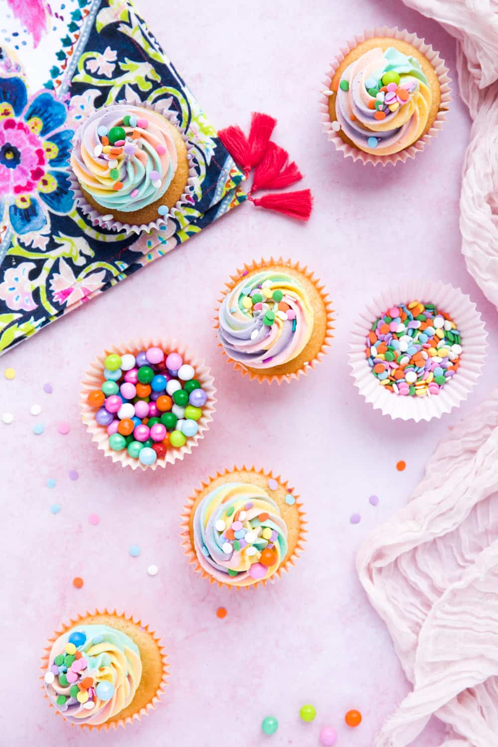 Overhead shot of cupcakes with rainbow coloured icing. There is a cupcake liner filled with round colourful sprinkles and a brightly coloured napkin. 