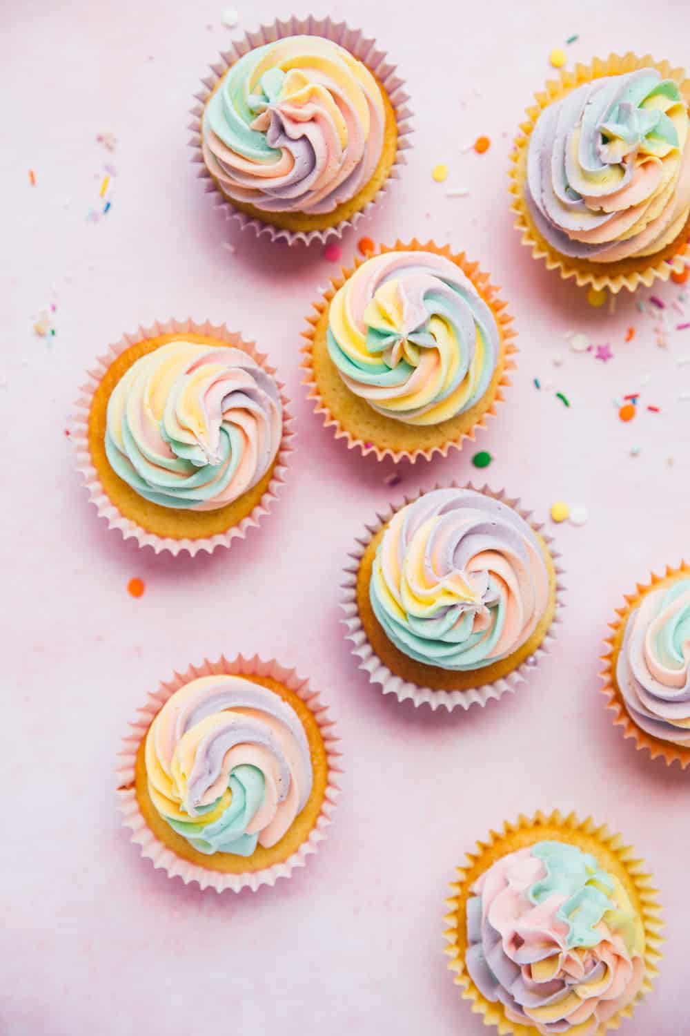 Overhead shot of cupcakes with rainbow coloured icing. There is a cupcake liner filled with round colourful sprinkles and a brightly coloured napkin. 
