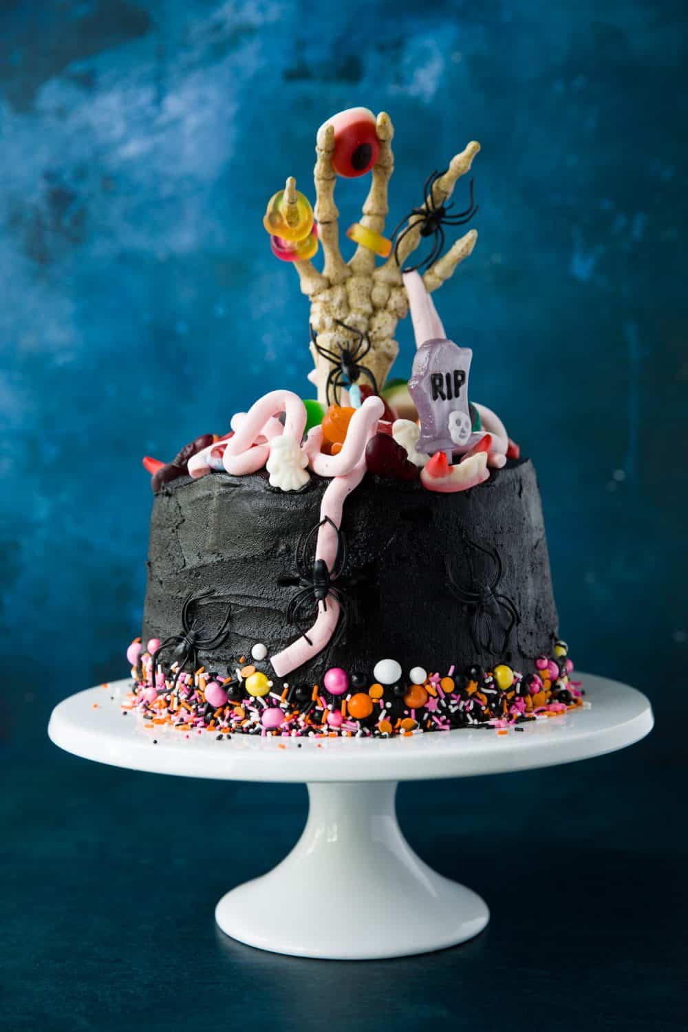 A 5 layer chocolate cake covered with black buttercream. There are colourful sprinkles around the bottom of the cake and a skeleton hand sticking out from the middle. The top of the cake is decorated with Halloween sweets.