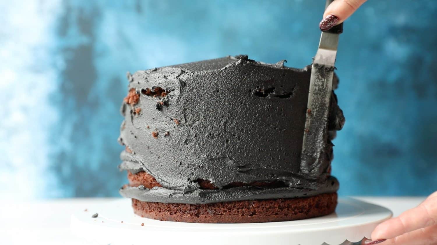 A five layer chocolate cake that has been covered in black buttercream icing. The icing is being spread over the cake using a spatula.