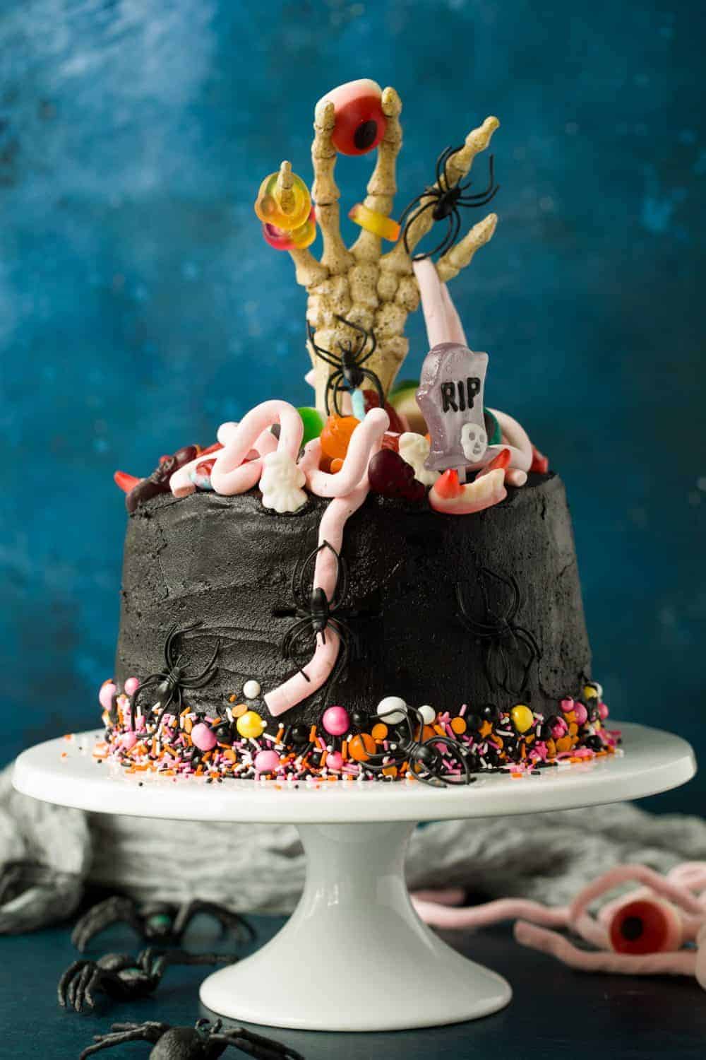 A 5 layer chocolate cake covered with black buttercream. There are colourful sprinkles around the bottom of the cake and a skeleton hand sticking out from the middle. The top of the cake is decorated with Halloween sweets.