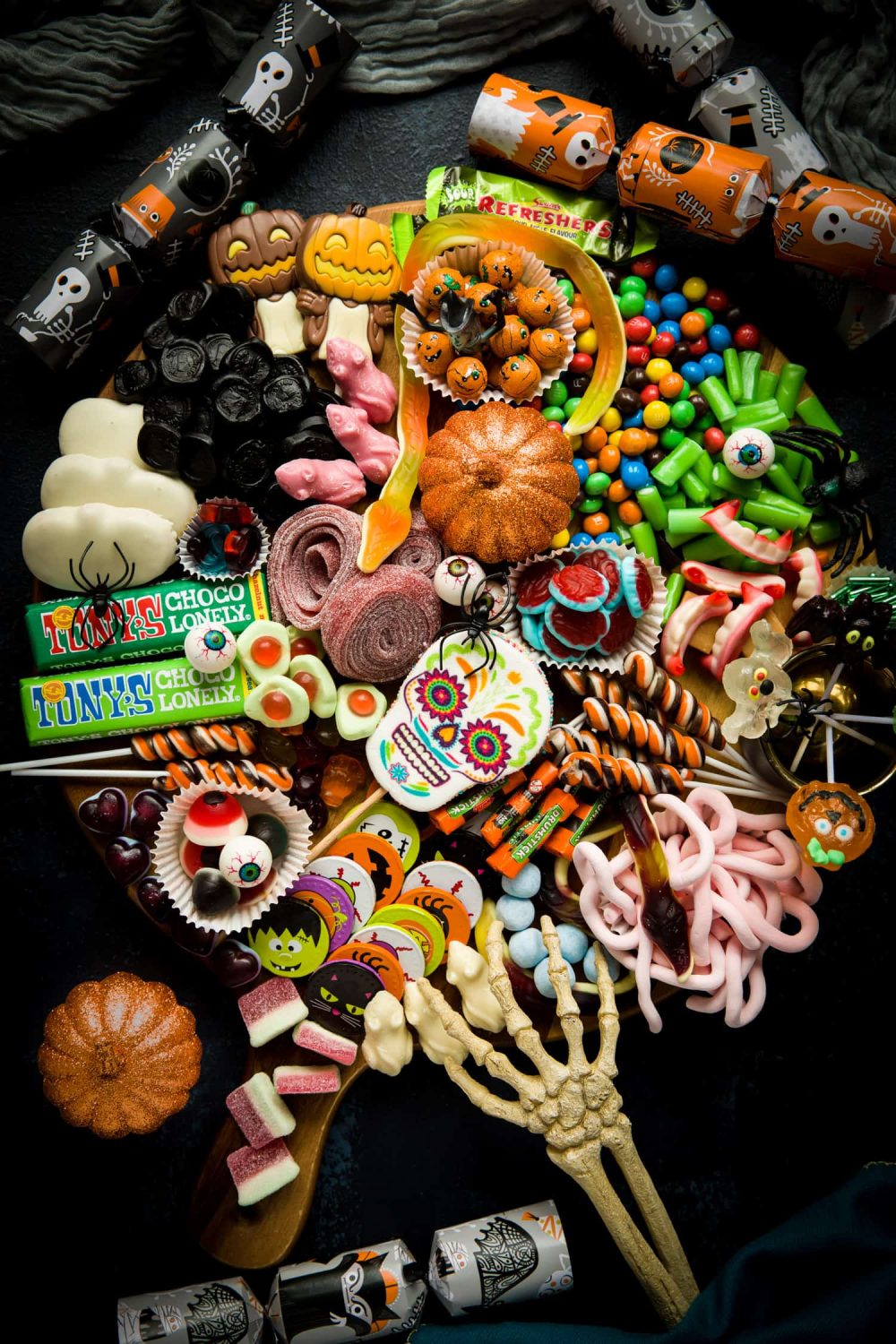 A Halloween Treat Platter filled with a wide variety of sweets, candy and chocolates. It is surrounded by halloween crackers and there is a skeleton hand reaching on to the tray. 