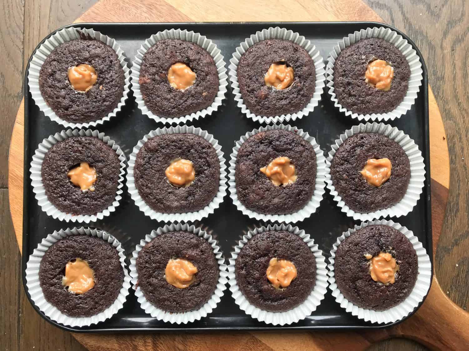 A tray of 12 chocolate cupcakes with holes in the centre. Each hole has been filled with caramel sauce.