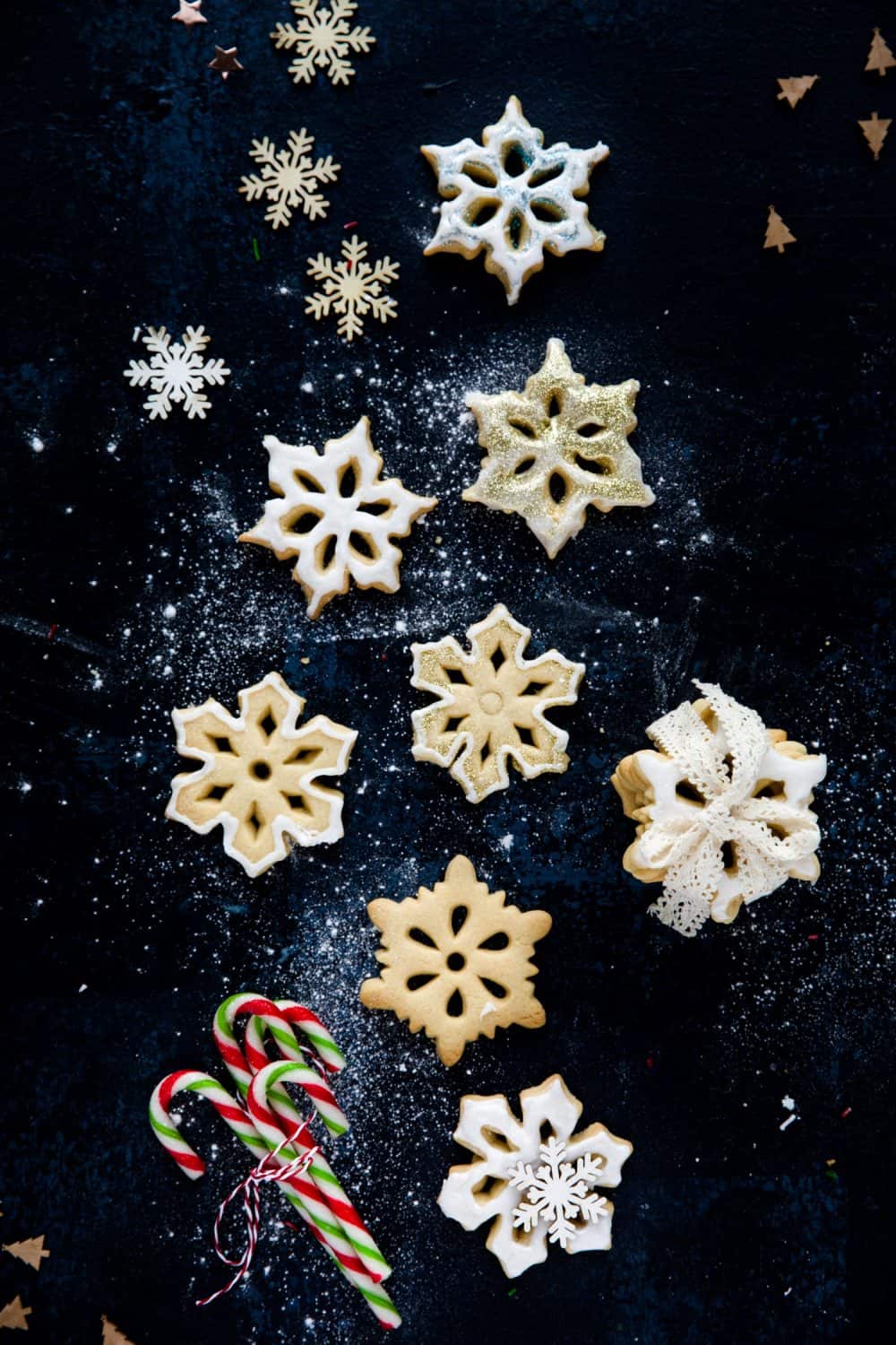 Overhead view of 7 snowflake biscuits on a dark blue background. There is a stack of biscuits to the right and at the bottom of the image a bundle of candy canes. 