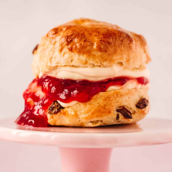 A small pink cake stand with a fruit scone on top. There is jam and clotted cream oozing out and dripping down the left side of the scone.
