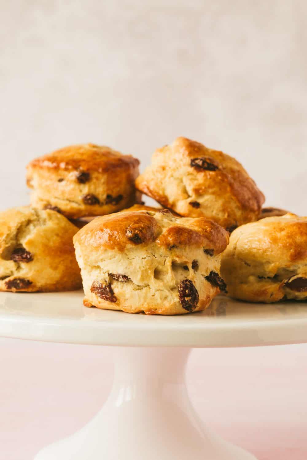 Side view of 6 fruit scones on a white cake stand. There are visible sultanas protruding from the scones and they are golden brown in colour. 