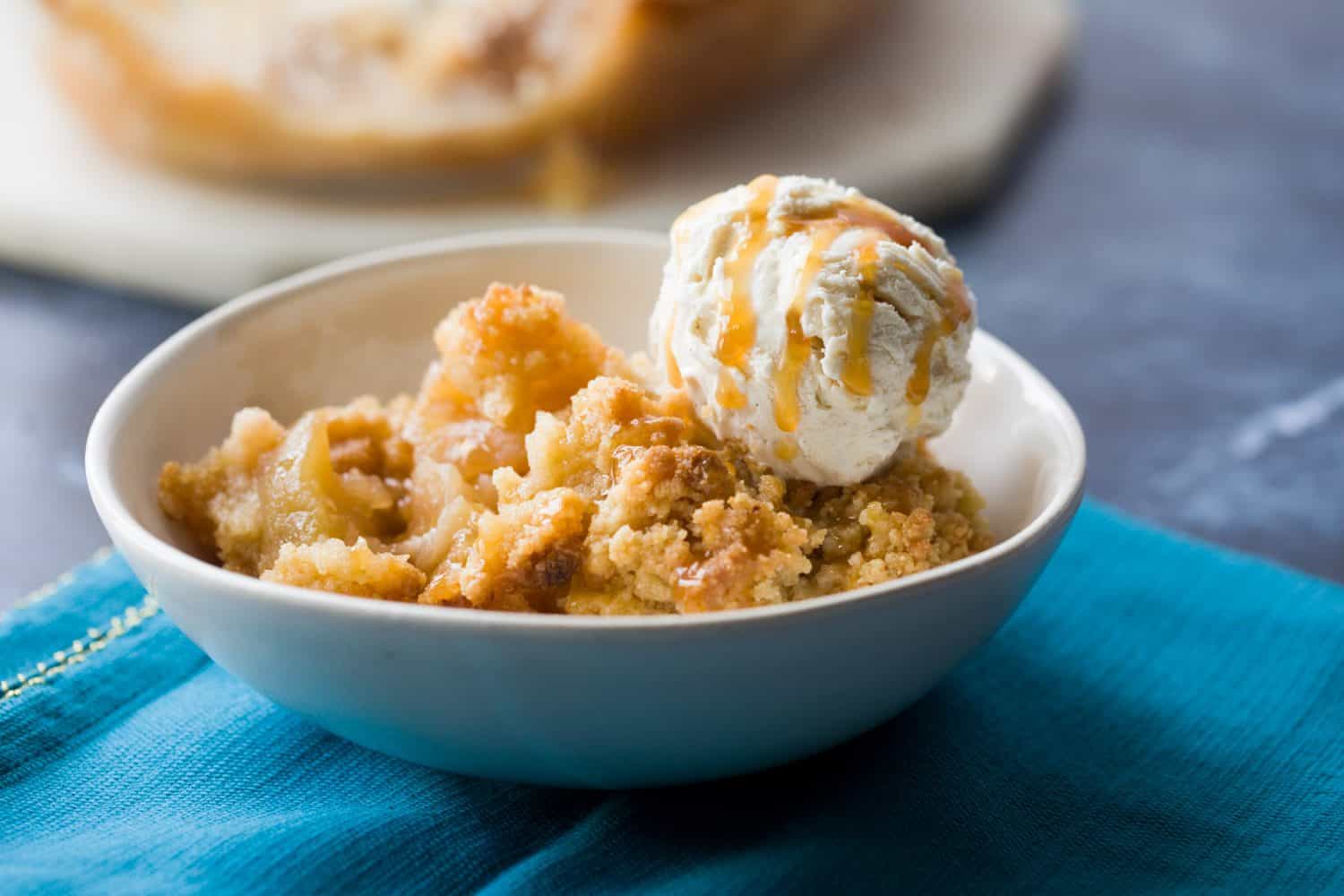 A white bowl with a portion of apple crumble. There is a scoop of ice cream on top and the dessert has been drizzled with caramel sauce. 
