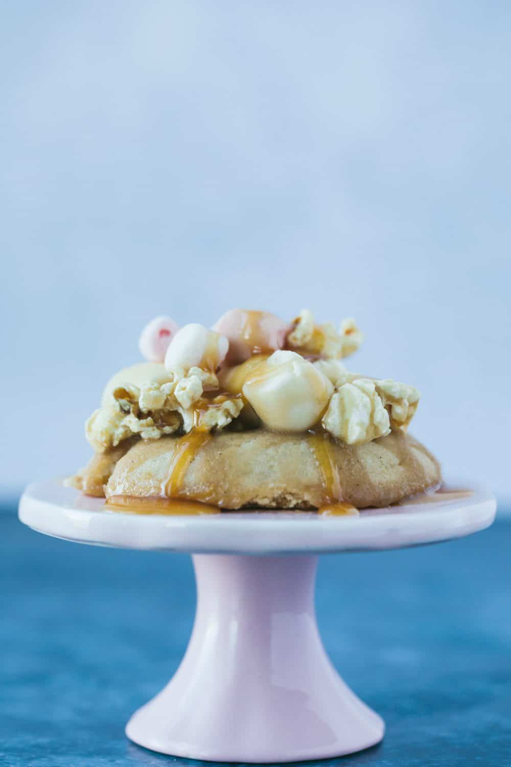 A small pink cake stand with a cookie that has popcorn and marshmallows on top. The cookie has been drizzled with caramel. 