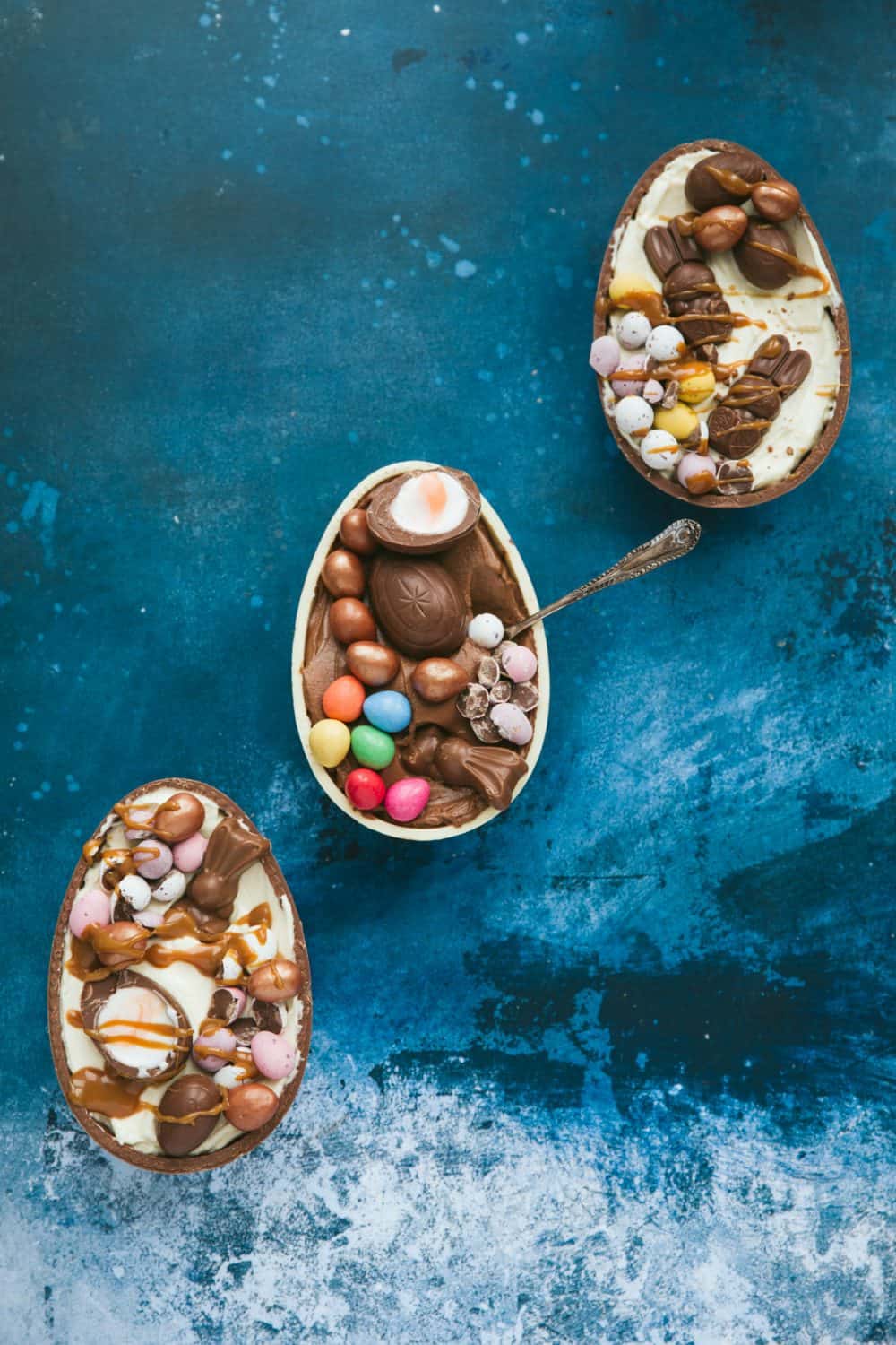 A dark blue background with three halves of Easter eggs each filled with cheesecake and topped with Easter chocolate. 