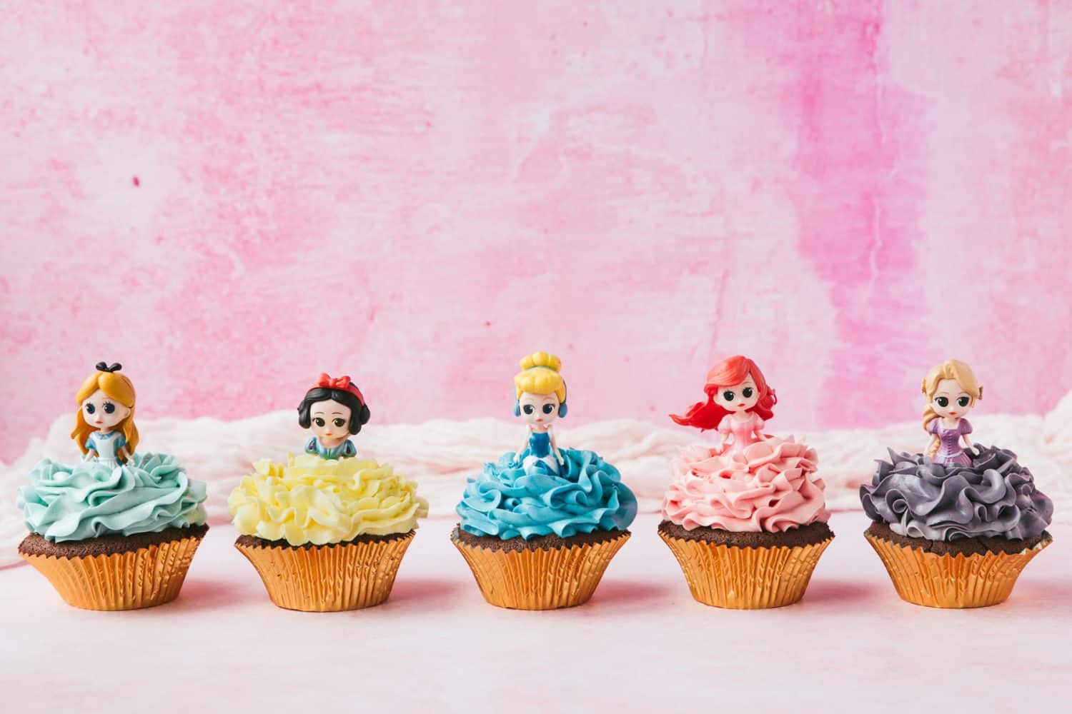 A row of Disney Princess Cupcakes. Alice in Wonderland, Snow White, Cinderella, Ariel and Rapunzel with coloured frosting. 