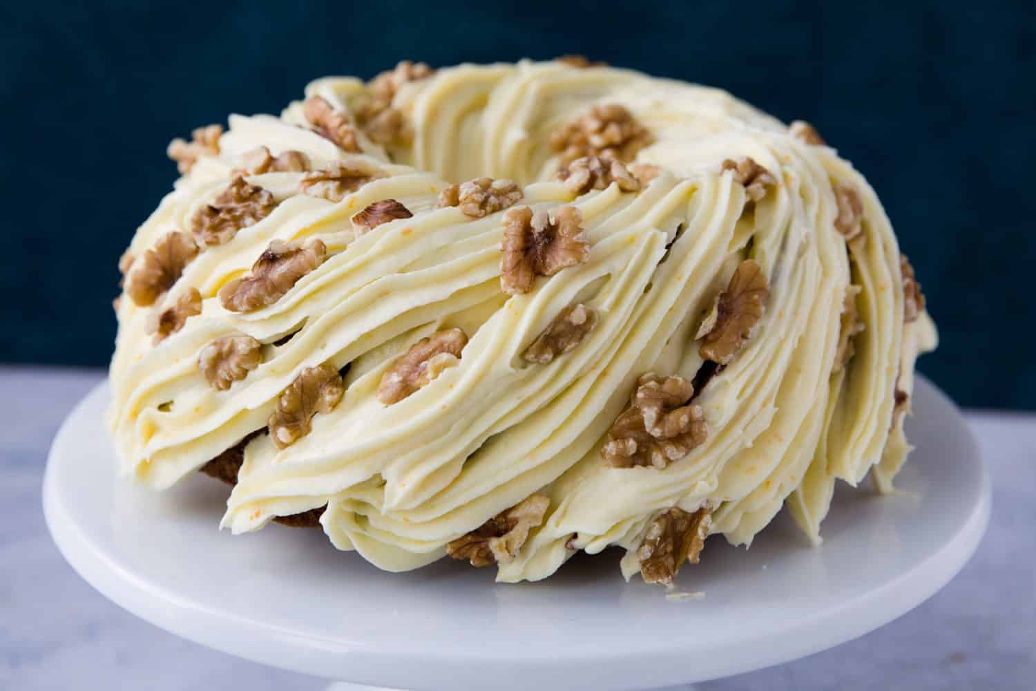 A carrot cake bundt on a white cake stand. There is frosting all over the cake and whole walnuts have been added for decoration. 