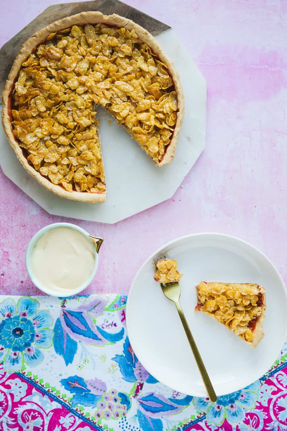 A cornflake tart with a slice cut out and a piece on a white plate beside a small pot of custard. 