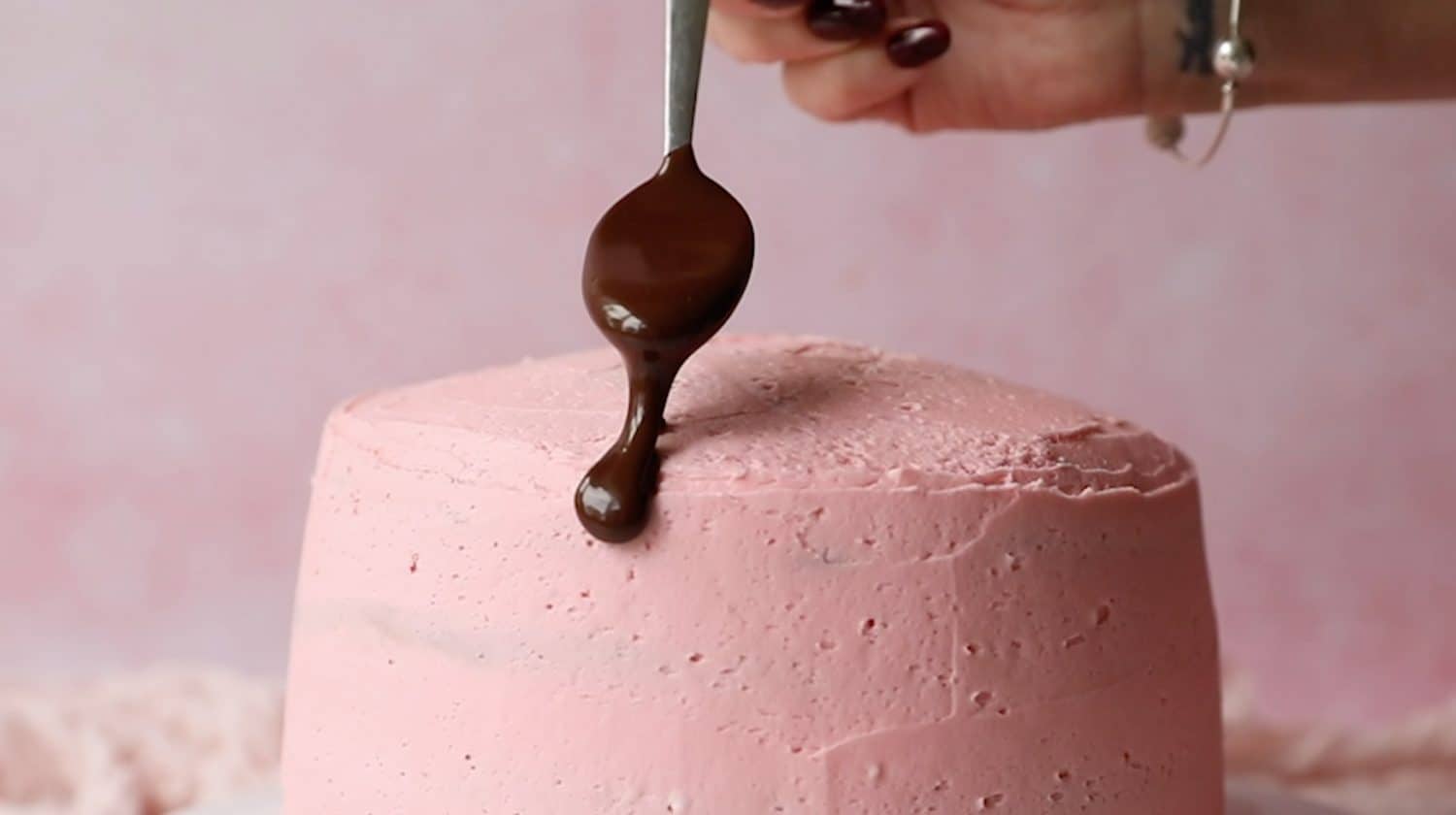 A hand holding a spoon of chocolate sauce that is being dripped on to a pink cake. 