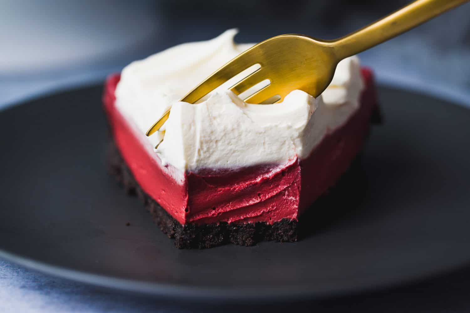 A slice of red cheesecake with a dark crust and cream topping. 