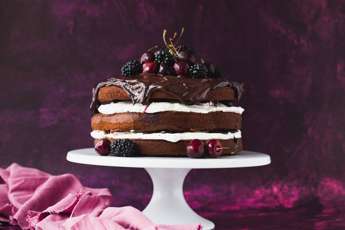 A black forest cake sitting on a white cake stand. 