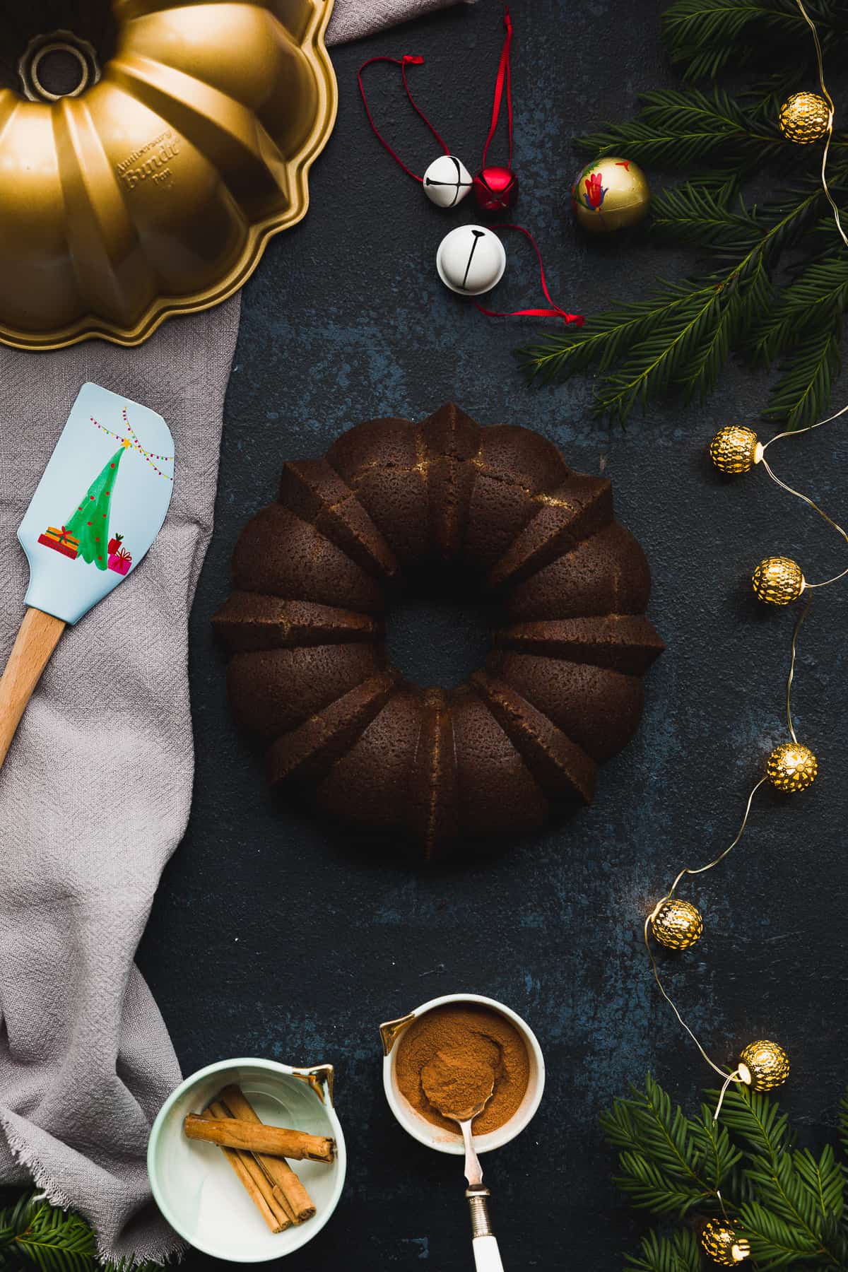 A black board with a gold bundt tin in the corner. In the centre is a dark coloured gingerbread bundt cake surrounded by christmas decorations and lights. 