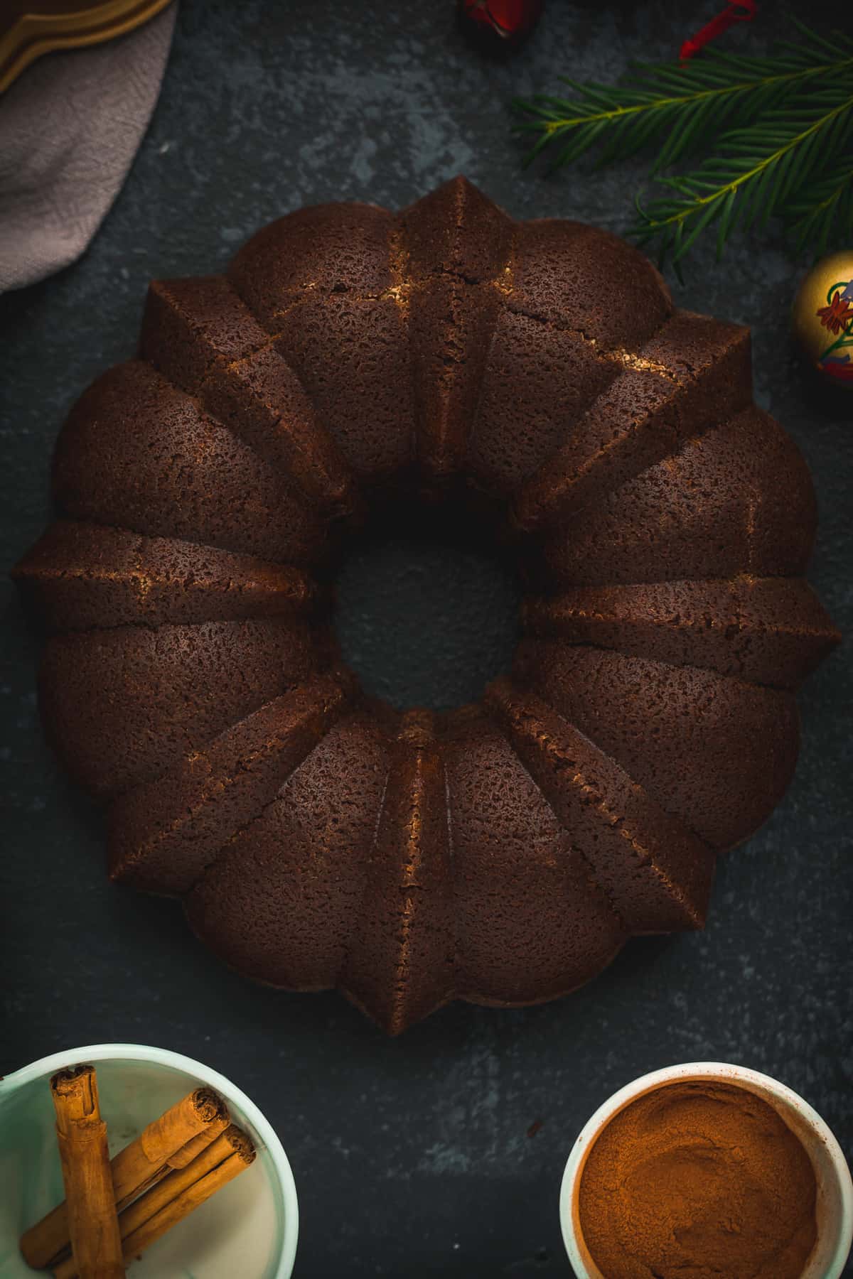 A ginger cake on a very dark background. There is a bowl of ground ginger next to it and another bowl with cinnamon sticks. 