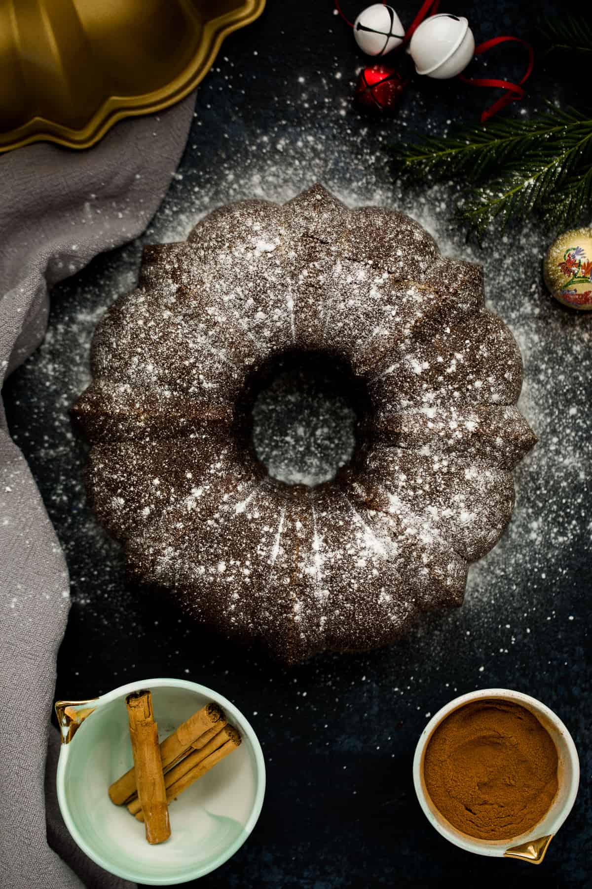 A black board with a gold bundt tin in the corner. In the centre is ginger cake in a circular shape, it has been dusted with icing sugar. It's surrounded by christmas decorations and lights. 