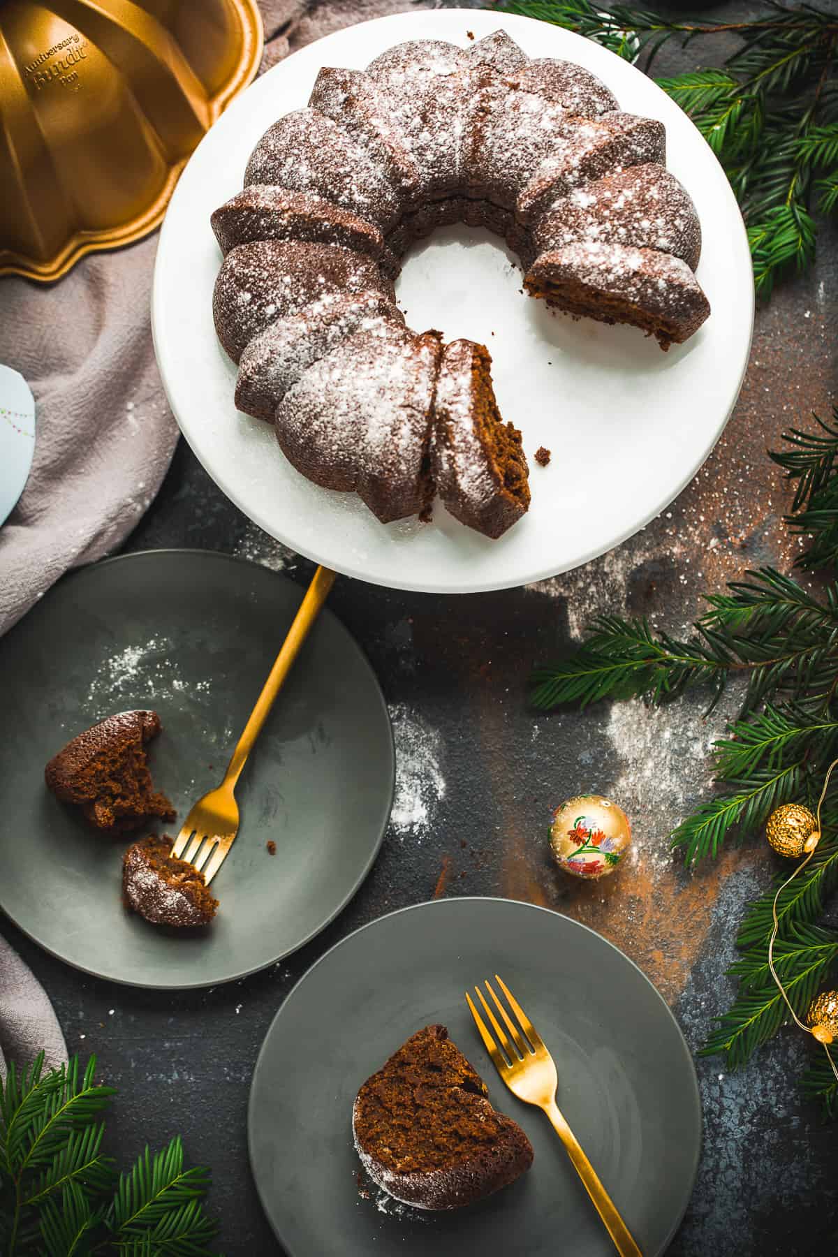 Overhead view of a bundt cake on a white platter. Two pieces have been cut out and they have been placed onto dark grey plates with gold coloured forks. The scene is surrounded by Christmas decorations and looks very festive. 