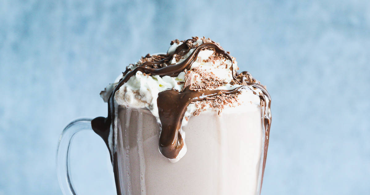 A hot chocolate made from Galaxy powder, milk and Baileys. There is cream on top and chocolate sauce is dripping down the side of a glass mug. 
