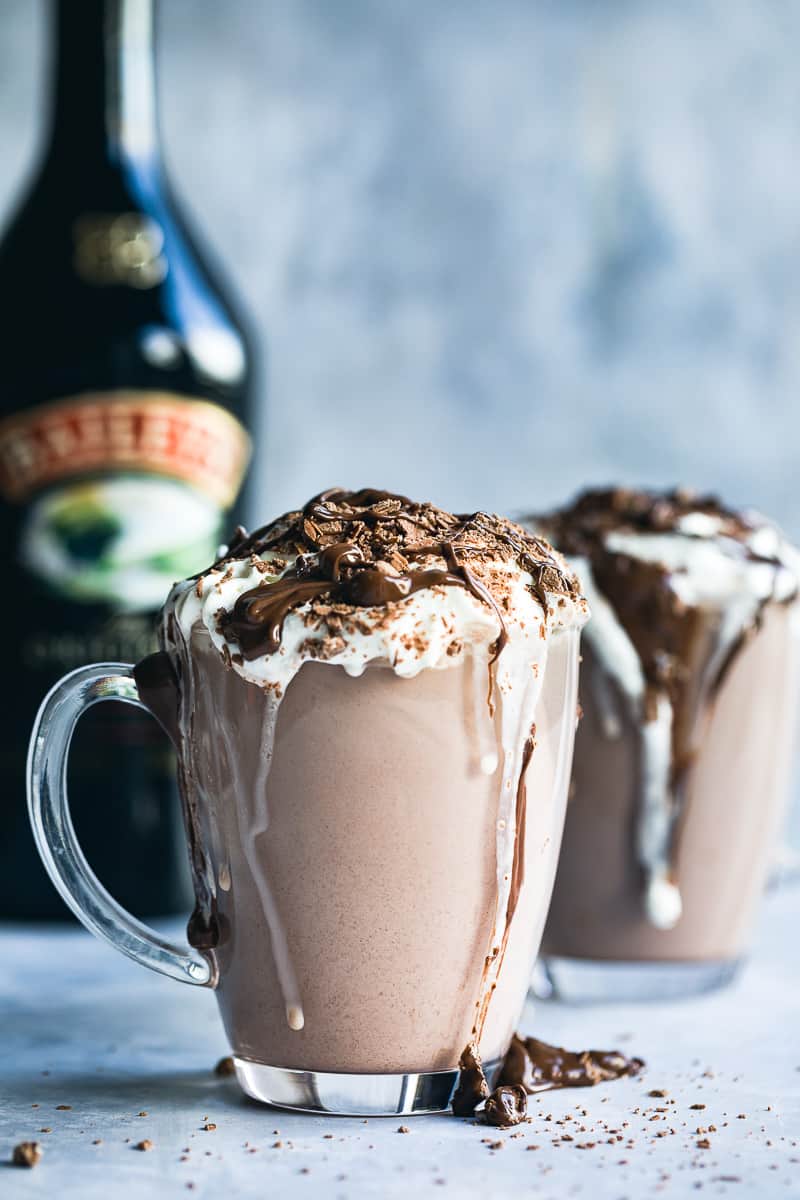 A glass mug filled with Baileys hot chocolate and topped with whipped cream a crumbled flake chocolate bar and a drizzle of chocolate sauce. 