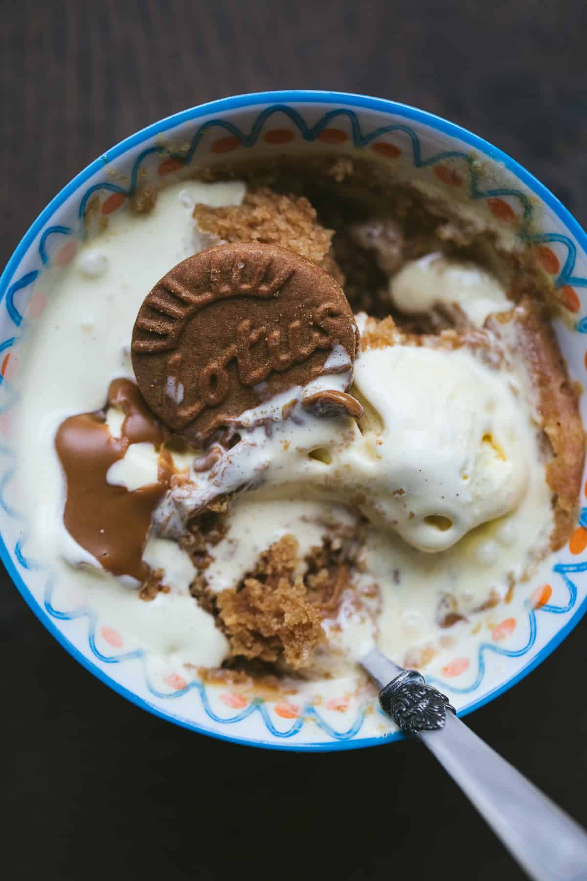 A blue mug filled with a mug cake and topped with ice cream that has melted. The ice cream has had warm Biscoff spread poured o to it and there is a round Biscoff Creme biscuit poking out of the top. 