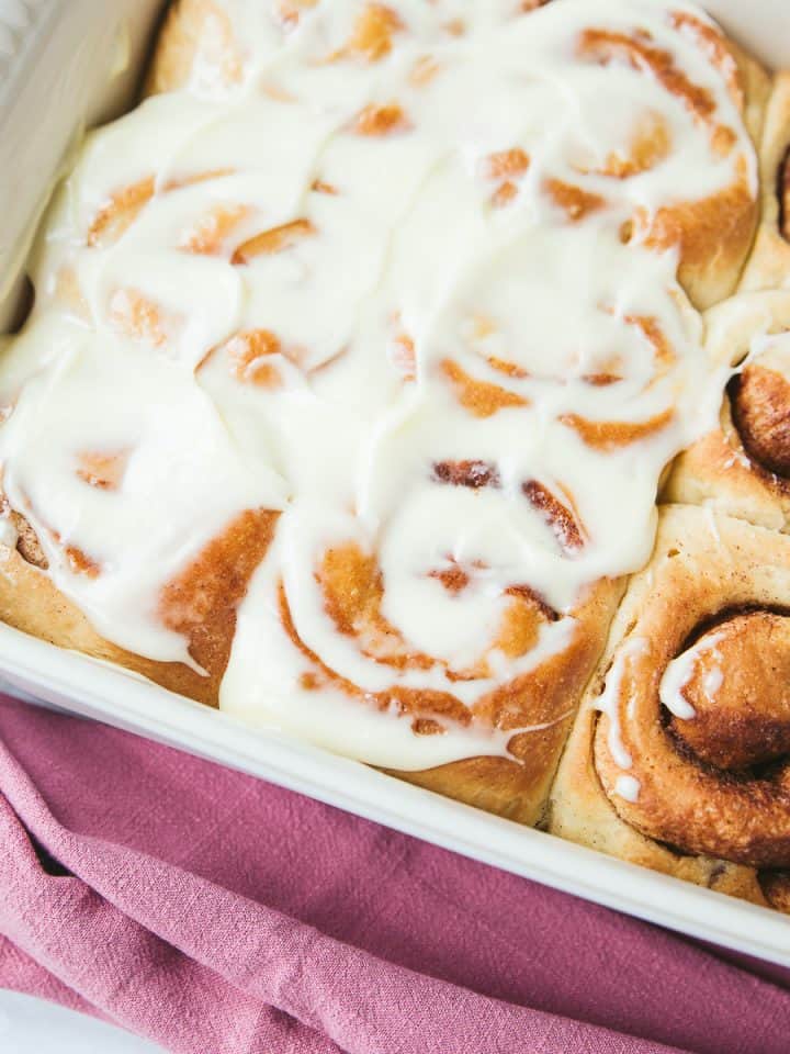A white baking dish containing Cinnamon Rolls, half have been spread with cream cheese frosting and the other half have been left plain.