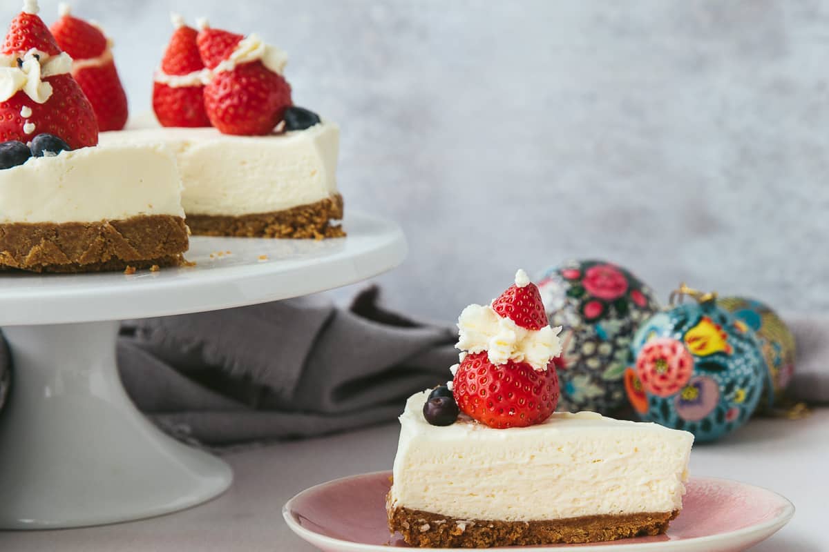 A slice of cheesecake on a pink plate surrounded by a festive scene. 