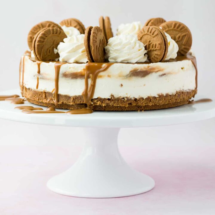 A cheesecake drizzled with biscoff spread and topped with cream and biscuits on a white cake stand.