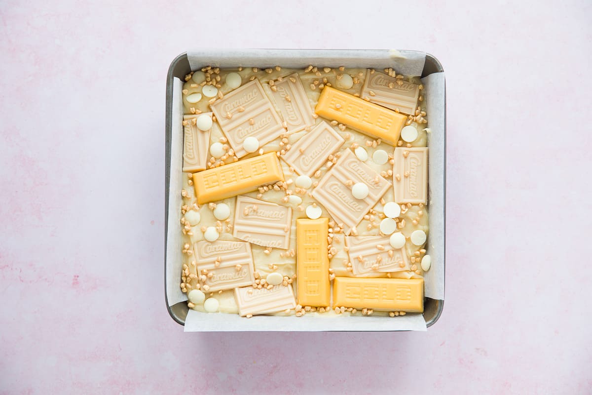 A square baking tin with a white chocolate blondie mixture. Gold bars and caramac bars have been added to the top. 