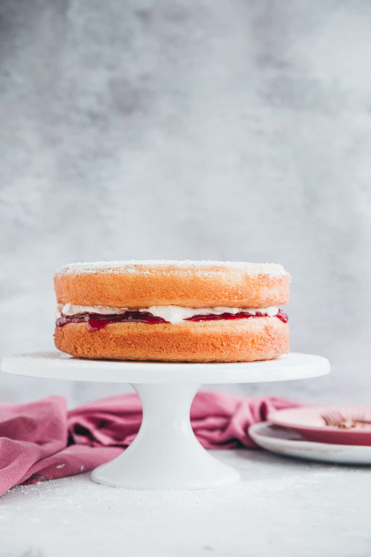 A two layer Victoria Sponge filled with freshly whipped cream and strawberry jam. The cake is on top of a white cake platter against a pale grey background. 