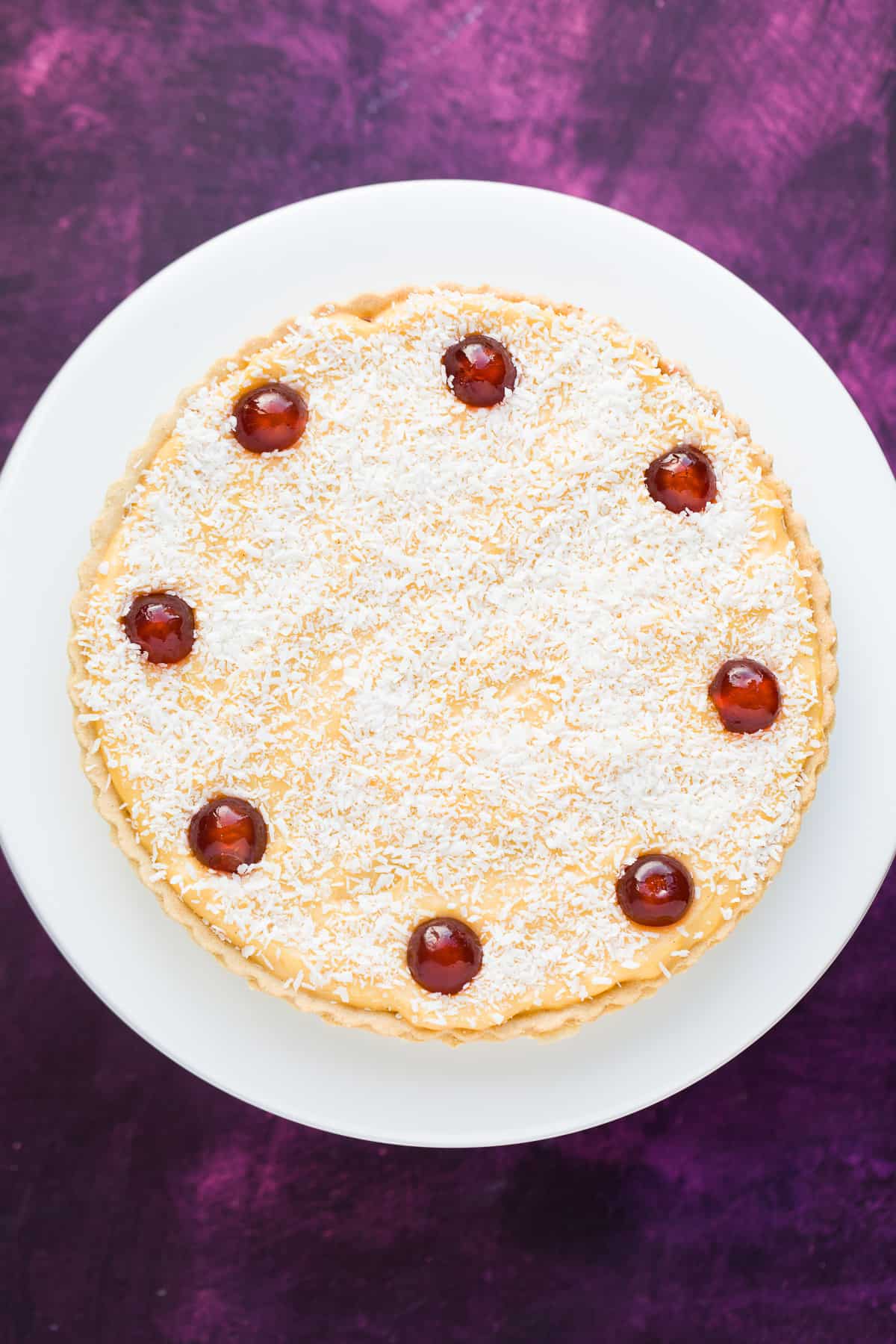 A Manchester tart with sweet pastry, raspberry jam, sliced banana, homemade custard and topped with desiccated coconut and maraschino cherries. 