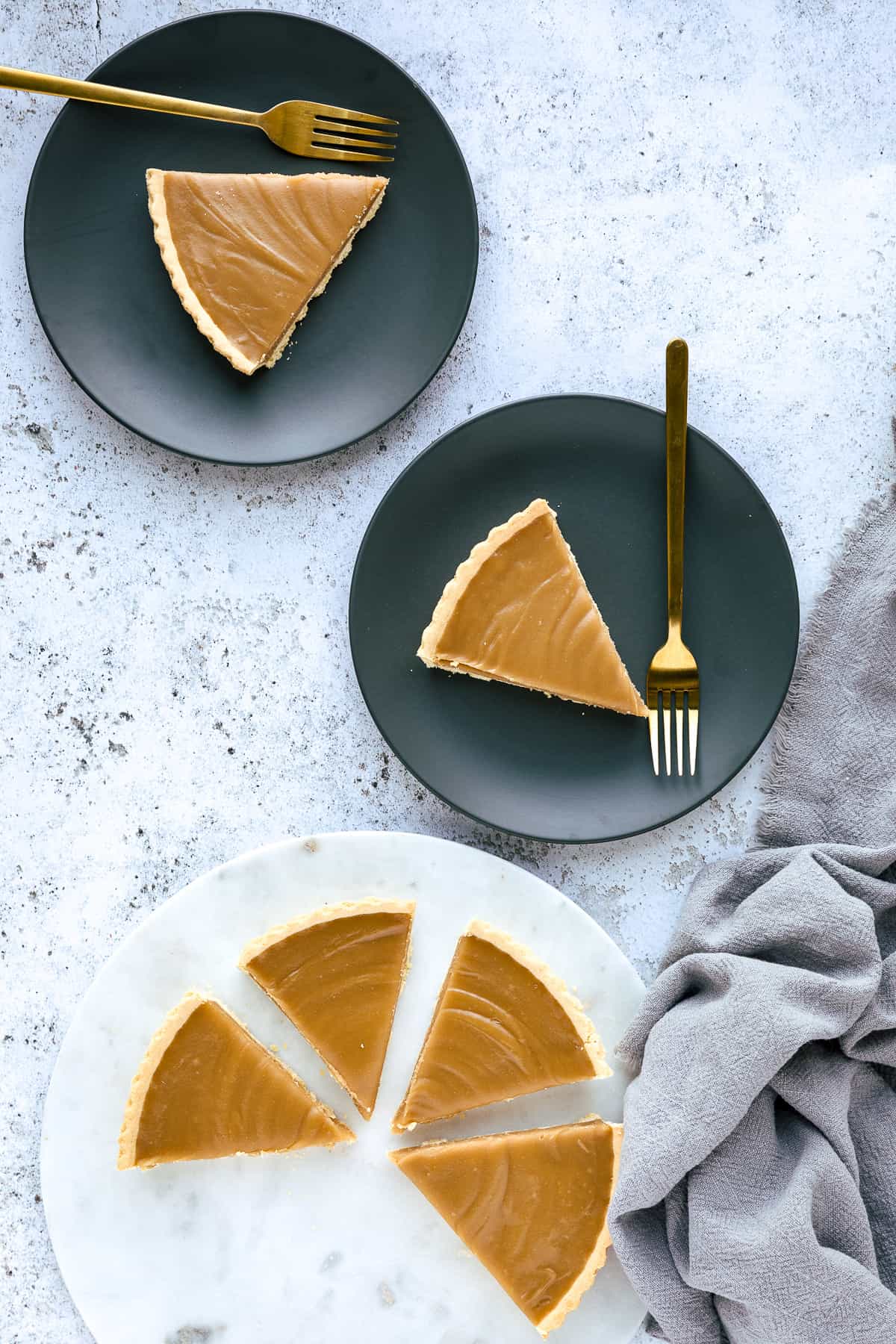A Butterscotch Tart that has been sliced, there are two pieces on small grey plates and the rest is on a white marble chopping board. 