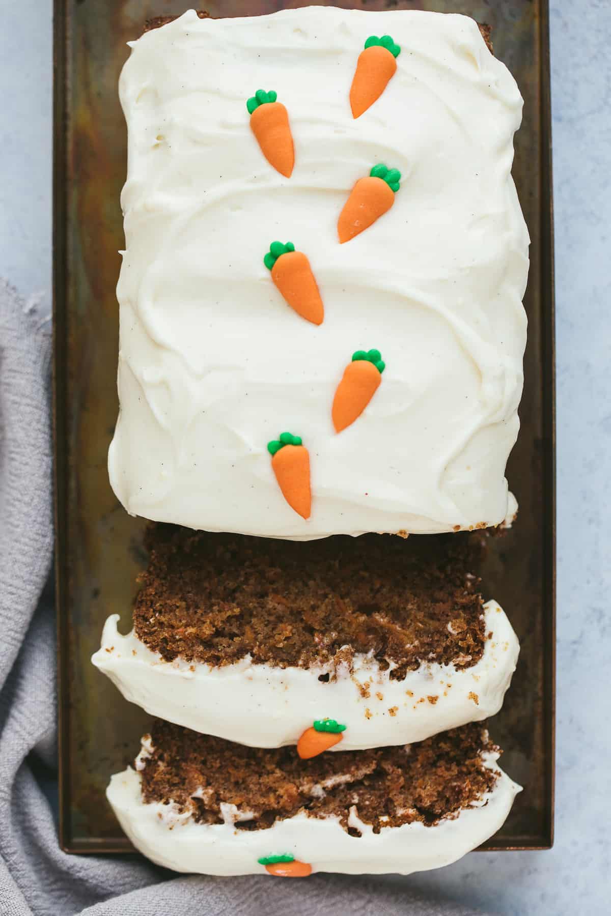 A carrot cake loaf that has two slices cut away. There is a cream cheese frosting on top and tiny fondant carrots. 