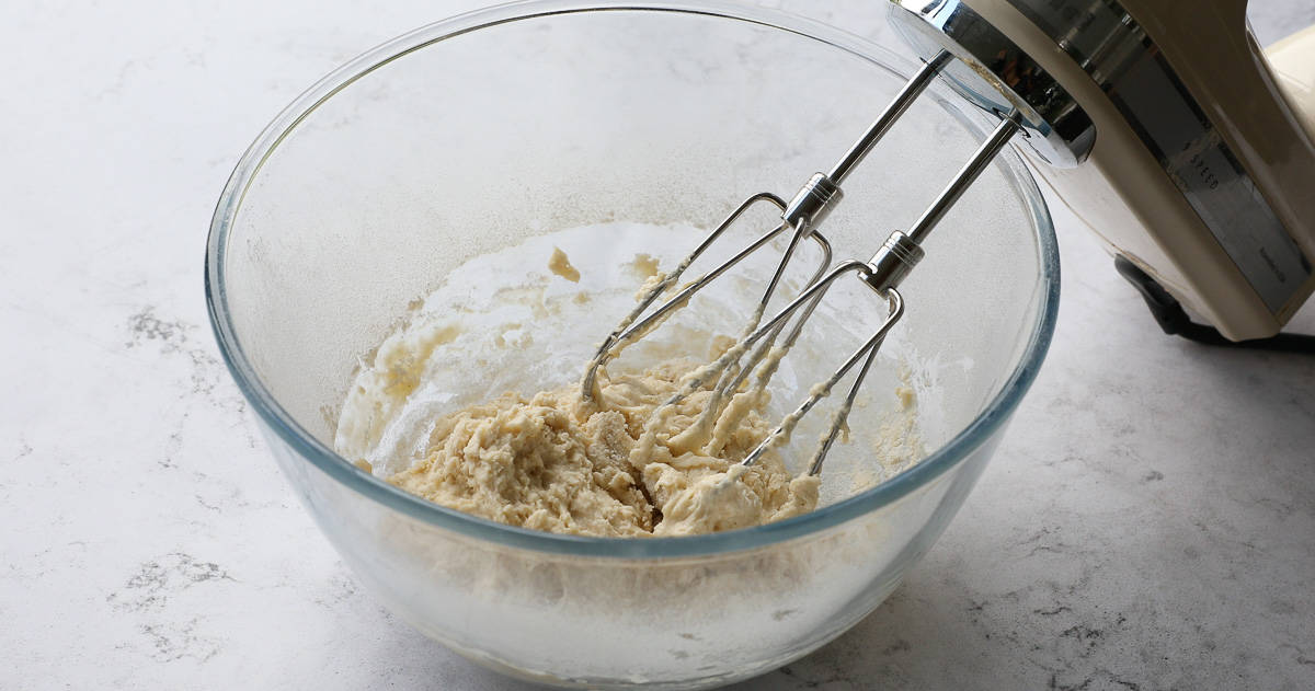 A glass mixing bowl with egg mixture and flour that has been beaten together with a handheld electric whisk. 