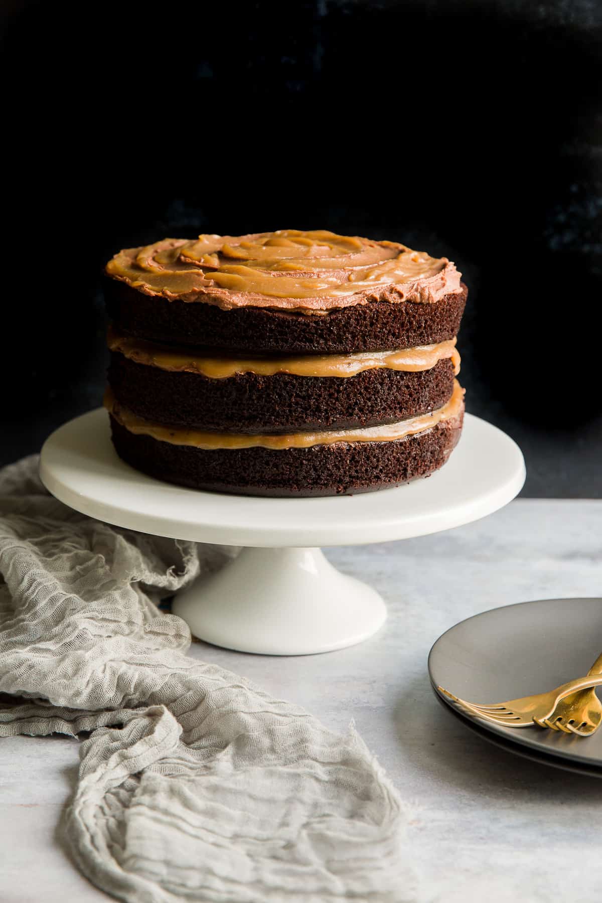 A three-layer chocolate salted caramel cake on a white cake stand set against a dark background. 